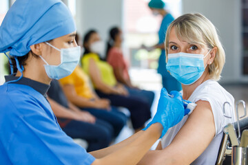 Asian female doctor wears face mask and blue hospital uniform injecting vaccine to Caucasian senior woman at ward working desk with equipment while other patients wait in queue in blurred background