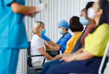 Doctor wears face mask injecting vaccine to Caucasian woman while nurse getting personal information from female patients sitting waiting in queue to covid 19 vaccinating in blurred foreground