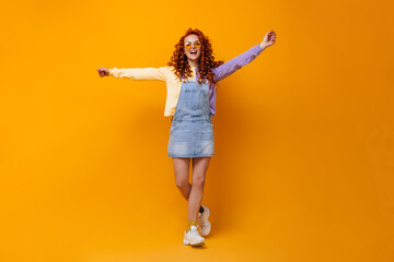 Fototapeta na wymiar Curly woman in sunglasses, denim sundress and sweater having fun on orange background