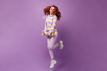 Naughty girl in purple glasses, light trousers and sweatshirt holds skateboard and jumps on isolated background