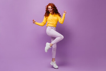 Fototapeta na wymiar Slender girl in light pants and yellow sweater dances against lilac background