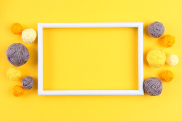 A white empty frame and a lot of gray and yellow balls of yarn for knitting or crocheting on a yellow background.The concept of manual labor, crafts, hobbies.Top view.Flatlаy.Copyspace.