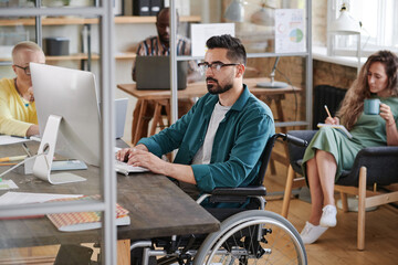 Serious young businessman in wheelchair sitting at the table and typing on computer he working at office with his colleagues