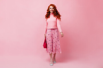 Red-haired girl in pink skirt and sweater posing with bright bag on isolated background