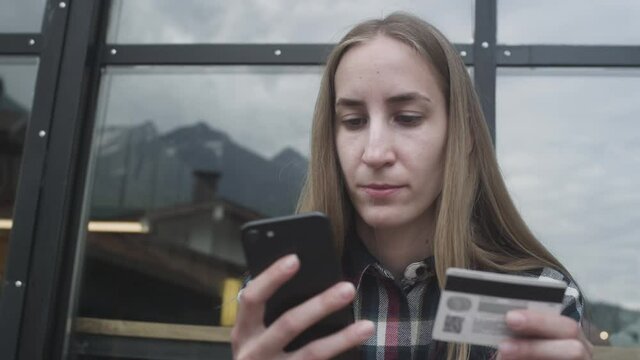 Young beautiful girl uses a smartphone and a bank card in a cafe against the backdrop of mountains. She makes purchases, buys plane tickets for travel. Downshifting during a pandemic and quarantine.