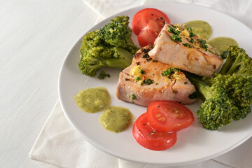 Healthy meal with fried tuna fillet, broccoli, tomatoes, parsley and green lemon sauce on a white plate and a light table, copy space, selected focus
