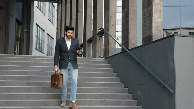 Full length portrait of arabian man walking down on stairs with modern smartphone in one hand and leather brown suitcase in another. Concept of business, success and confidence. Arabian man