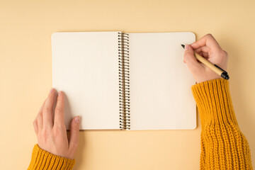 First person top view photo of female hands in yellow knitted sweater writing in spiral notebook on isolated pastel orange background with blank space