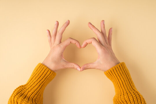 First person photo of female hands in yellow pullover making heart with fingers on isolated light orange background