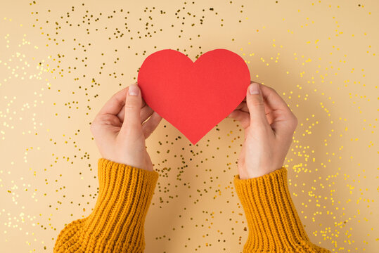 First person top view photo of female hands in yellow pullover holding red paper heart over scattered golden sequins on isolated light orange background with copyspace