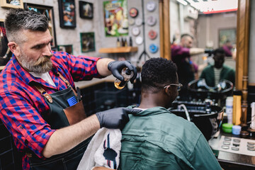 Young African man visiting hairstylist in barber shop. Professional hairdresser cut hair with electric shearer machine.