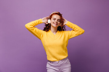 Cute girl in sweater listens to music in white headphones and dances on purple background