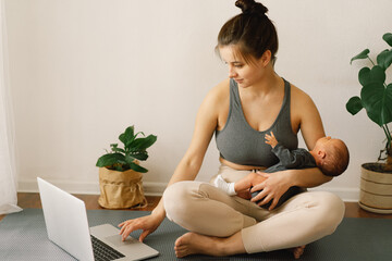 Mother holds her newborn baby son and works at the computer at home.