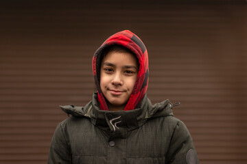 Young boy in a hoodie of a sweatshirt and a jacket against background of brown blinds on the street
