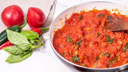 Making homemade Mexican hot tomato sauce in a skillet with ingredients on the table - chili peppers, jalapenos, tomatoes and basil