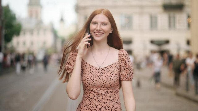 Young beautiful red-haired woman with nice Freckles walking down the city street and talking on the phone. Girl smiling and communicating with friends