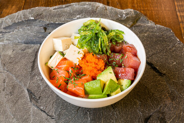 Poke bowl with fish roe, salmon, red tuna, green avocado, white tofu and guacamole seaweed with sesame seeds on gray stone background