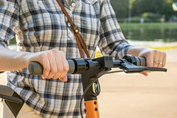 Woman in a checkered shirt holds the wheel of an electric scooter in a city park. Close-up of hands on the wheel of a scooter. City electric scooter rental service - kick sharing.