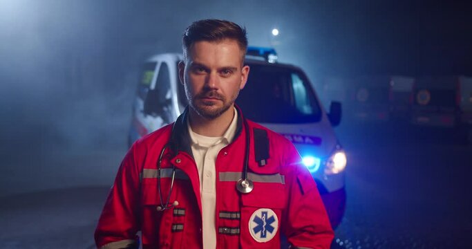 Portrait of Caucasian handsome young male paramedic in red uniform looking to camera and standing outdoors. Ambulance car on background. Attractive serious male doctor at night shift.