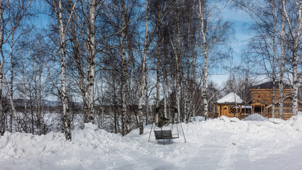 Fototapeta na wymiar Winter Siberian Landscape. Among the snowdrifts are wooden swing bench. A single-storey houses from unpainted logs are visible. Behind Birch Grove. Naked branches and trunks of trees against blue sky