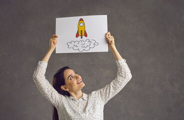 Joyful smiling woman holding paper banner with cartoon toy spaceship. Kindergarten nursery preschool school teacher showing space rocket picture. Innovation, success, education, business boost concept