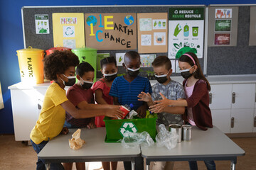 Group of diverse students wearing face masks putting recyclable plastic items in tray at school