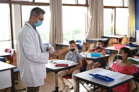 Caucasian male doctor wearing face mask showing how to use hand sanitizer to students at school