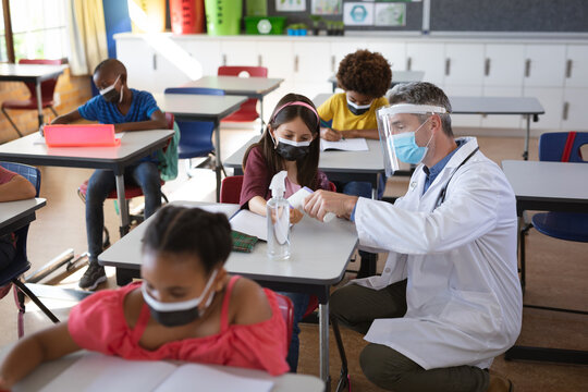 Caucasian male doctor wearing face shield measuring temperature of caucasian girl at school