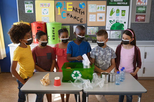 Group of diverse students wearing face masks putting recyclable plastic items in tray at school - Powered by Adobe