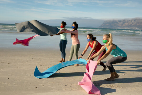 Group of diverse female friends wearing face masks putting down mats, practicing yoga, at the beach