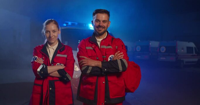 Portrait of happy Caucasian young man and woman paramedics at ambulance. Male and female doctors in red uniforms smiling at camera. Physicians and colleagues at night during coronavirus pandemic.