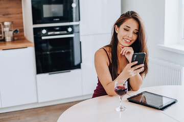 attractive Asian woman drinks wine using a mobile phone and using a tablet in the kitchen at home