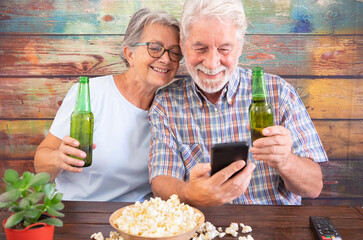 Smiling senior couple sitting at pub drinking beer and eating popcorn. Watching at mobile phone, wooden background