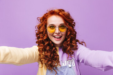 Fototapeta premium Cheerful woman with red hair laughs and makes selfie on lilac background