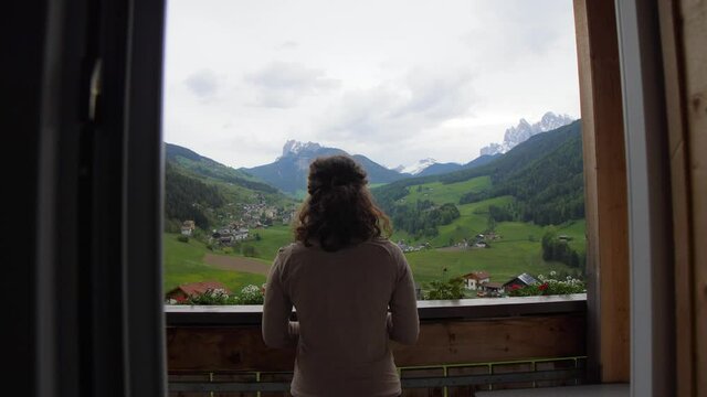 Silhouette of a girl going on a balcony with mountain view in the Italian Alps. A young woman traveler in an alpine hut enjoying the panorama of the Val di Funes valley. Dolomites, Italy.