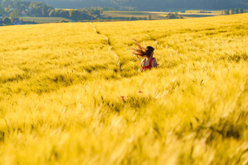 Pretty teenager girl with long hair enjoying nature running through yellow barley field in countryside at sunny summer day. Healthy holidays lifestyle.
