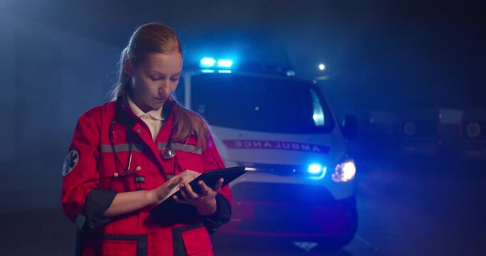 Caucasian young female paramedic in red uniform tapping and typing on screen of tablet computer. Woman medic filling in medical form on device at night outdoor. Emergency ambulance call.