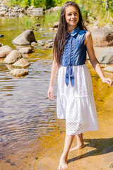 An attractive teenage girl in a summer dress stands on the shore of a reservoir with bare feet on a summer sunny day. 