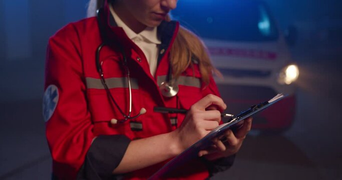 Close up of Caucasian female paramedic in red uniform writing with pen in hand in folder with medical cards and documents. Woman medic filling in medical form at night outdoor. Emergency.