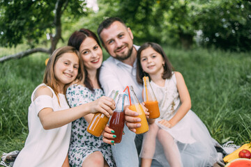 Smiling parents and their pretty daughters sitting on soft plaid outdoors and toasting with bottles of fresh juice. Family on picnic. Enjoyment concept. Focus on bottles with juice