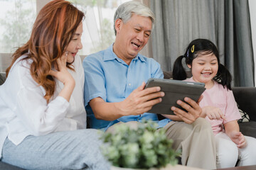 Obraz na płótnie Canvas Asian grandparents and granddaughter using tablet at home. Senior Chinese, grandpa and grandma happy spend family time relax with young girl checking social media, lying on sofa in living room concept