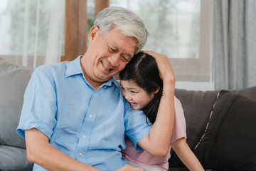 Asian grandfather talking with granddaughter at home. Senior Chinese, grandpa happy relax with young granddaughter girl using family time relax with young girl kid lying on sofa in living room.