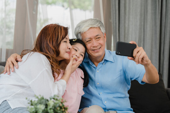 Asian grandparents selfie with granddaughter at home. Senior Chinese, grandpa and grandma happy spend family time relax using mobile phone with young girl kid lying on sofa in living room concept.