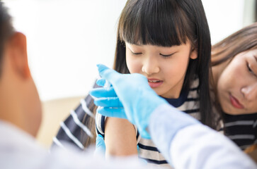 Asian  senior doctor wearing gloves and isolation mask is making a COVID-19 vaccination in the shoulder of child patient with her mother at hospital.