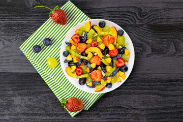 Salad in a white plate on a napkin made of fresh fruits from grapes, nectarine, kiwi and strawberries, blueberries, honeysuckle on a dark wooden background.Top view, copy  space