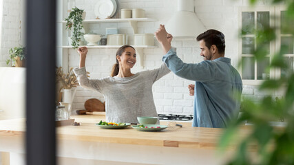 Carefree active millennial couple dancing together in cozy modern kitchen, listening to favourite music raise hands moving enjoy romantic date and happy relocation day to own rented fashionable house