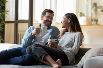 Calm relaxed couple hold cups rest on comfy sofa enjoy carefree talk while sit together on couch at...