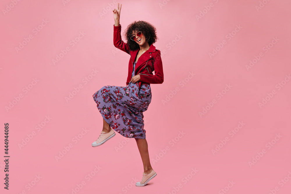 Wall mural Active girl in red jacket and flowered dress shows peace sign on pink background. Cool curly woman in floral skirt jumping on isolated backdrop