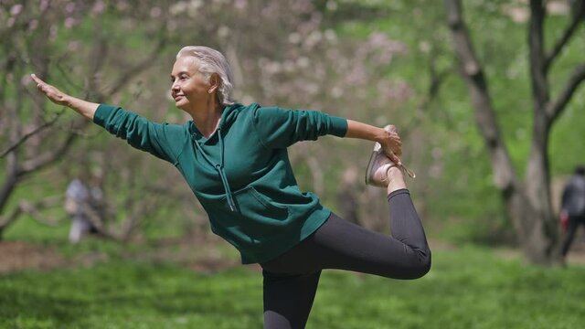 Happy senior sportswoman standing on one leg stretching muscles outdoors. Warmup exercise of positive confident fit Caucasian retiree in sunny spring summer park. Lifestyle and sport concept