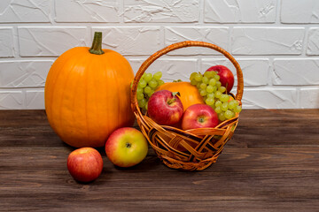 Orange pumpkins with apples and pears in a wicker basket on a brown wooden table against the background of a white brick wall. Autumn still life.